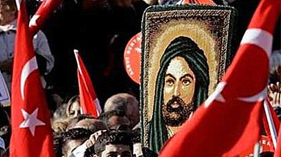 L'image montre une foule rassemblée lors d'une manifestation. Au centre, une personne tient une grande image représentant un personnage avec une longue barbe et un turban, entourée de drapeaux turcs. La foule semble engagée et énergique, avec des expressions variées sur les visages des participants.