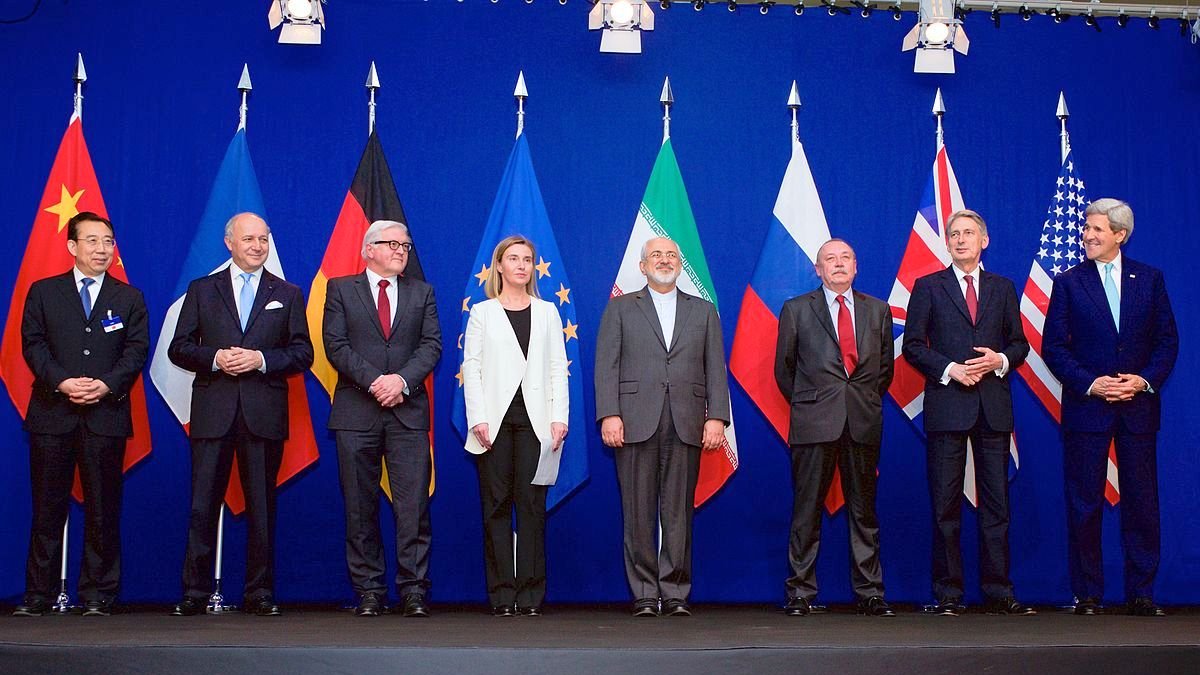 L'image montre un groupe de personnalités politiques posant ensemble devant un mur de drapeaux. On peut voir des représentants de plusieurs pays avec des drapeaux de la Chine, de l'Allemagne, de l'Union européenne, de l'Iran, de la Russie, du Royaume-Uni et des États-Unis en arrière-plan. Les personnes semblent discuter d'un sujet important et portent des costumes formels. L'ambiance est sérieuse, correspondant à un événement diplomatique.