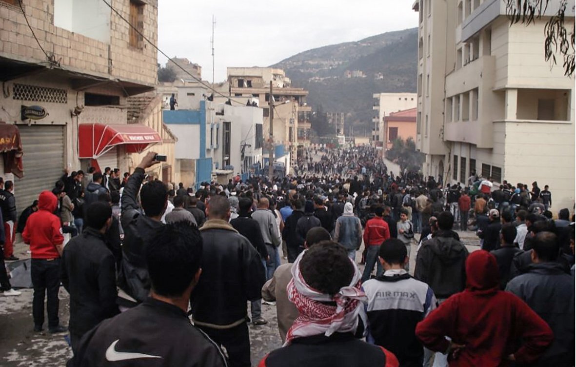 L'image montre une grande foule de personnes rassemblées dans une rue, entourée de bâtiments. Les gens semblent très nombreux, et certains portent des vêtements sombres ou des écharpes. L'atmosphère paraît tendue ou mobilisée, avec des manifestations potentielles. En arrière-plan, on aperçoit des constructions et des collines. L'ensemble donne une impression de mouvement collectif et d'unité.