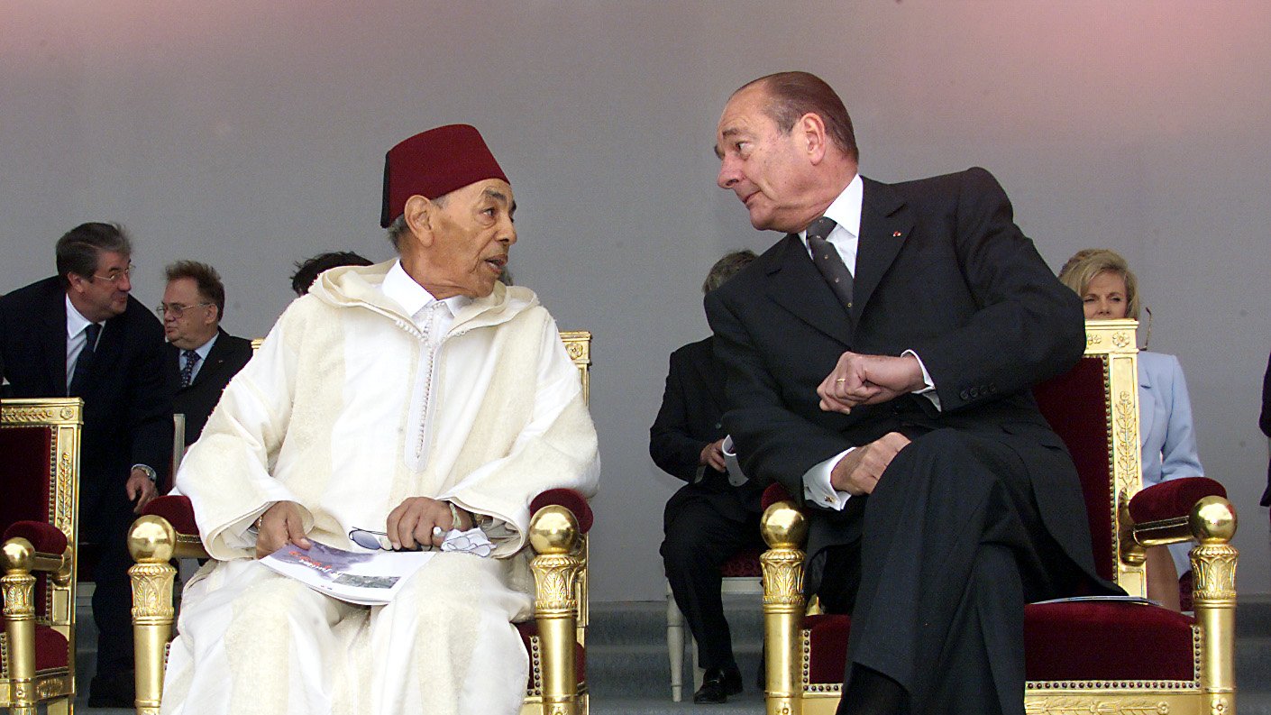 L'image montre deux hommes assis sur des chaises ornées, dans un cadre formel. L'un porte un habit traditionnel marocain avec un fez rouge, tandis que l'autre est vêtu d'un costume noir. Ils semblent engagés dans une conversation, se regardant avec attention. En arrière-plan, on aperçoit d'autres personnes, indiquant que cette scène a lieu lors d'un événement officiel. Leurs expressions semblent amicales et concentrées.