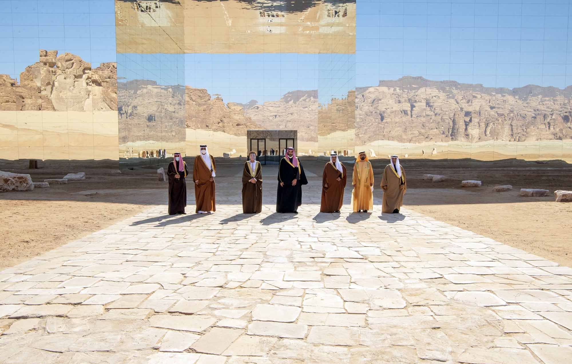 L'image montre un groupe d'individus en vêtements traditionnels, se tenant sur un chemin en pierre au milieu d'un paysage désertique. En arrière-plan, une grande structure réfléchissante fait un miroir du paysage environnant, créant un effet visuel intéressant. Les montagnes et le ciel sont également visibles, ajoutant une dimension naturelle à la scène. L'atmosphère semble solennelle et digne.