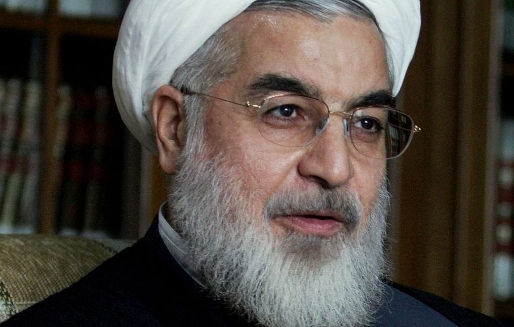 Je ne sais pas qui est représenté sur cette image. Cependant, je peux décrire des éléments généraux. L'image montre une personne portant un turban blanc, avec une barbe, des lunettes et des vêtements traditionnels. Il semble être en train de parler ou de s'exprimer de manière gestuelle. L'arrière-plan suggère un environnement intérieur, peut-être avec des bibliothèques ou des étagères.