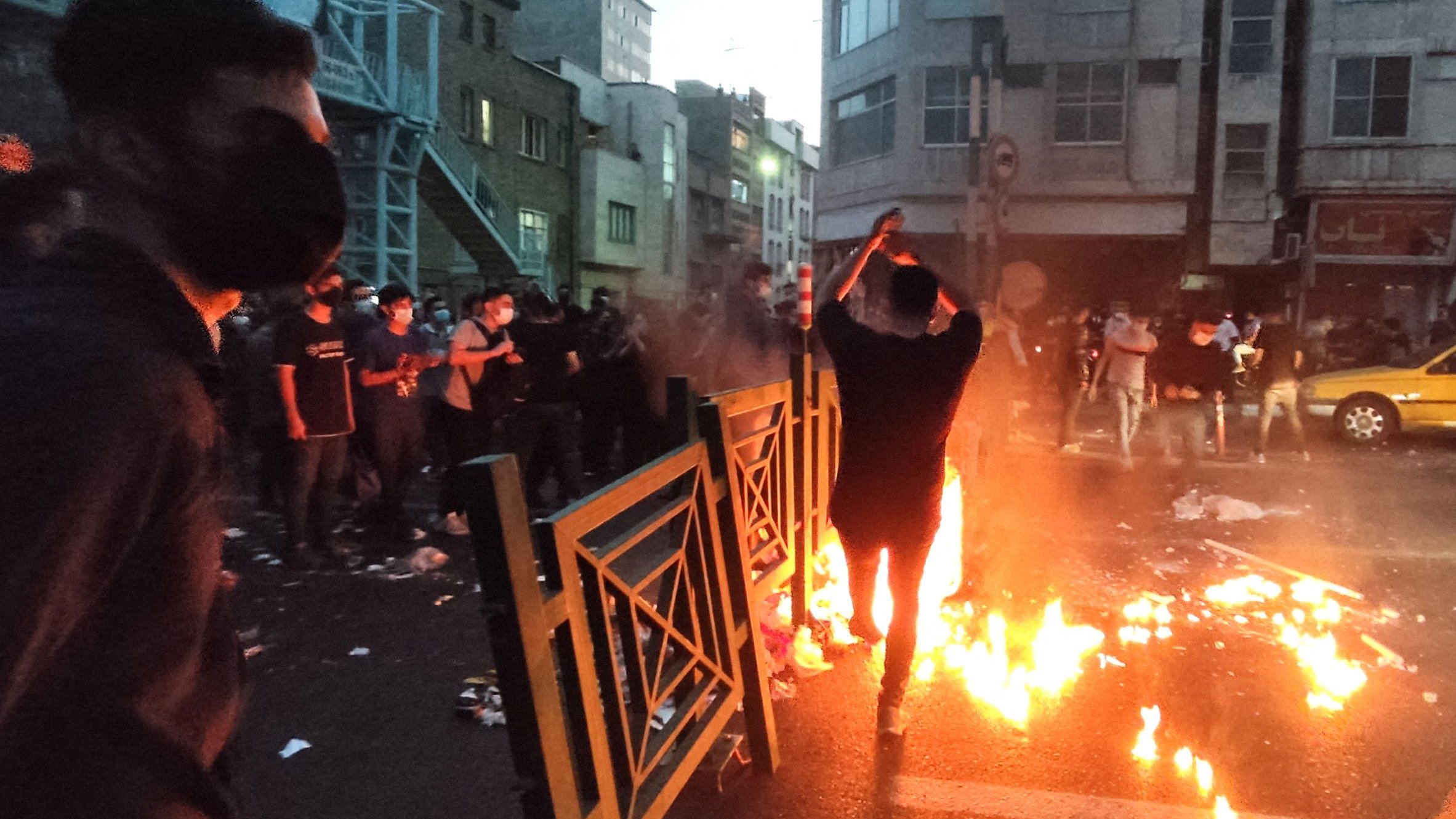 L'image montre une scène dynamique dans une rue urbaine, probablement lors d'une manifestation. On peut voir plusieurs personnes, dont l'une est au premier plan, se tenant près de flammes qui brûlent sur la route. L'atmosphère semble tendue, avec des barrières et des débris éparpillés. La lumière des flammes éclaire les visages des manifestants, qui portent des masques. En arrière-plan, on aperçoit des bâtiments urbains et une lumière tamisée, ce qui donne une impression d'urgence et de mouvement.