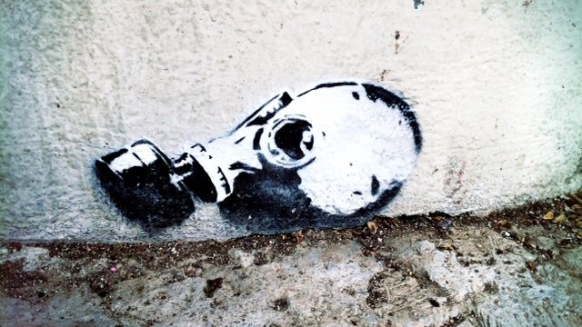 L'image montre un graffiti représentant une tête de mort portant un masque à gaz. Ce dessin est peint en noir et blanc et est appliqué sur un mur, près du sol. Le contraste entre le noir et le blanc met en évidence les contours du masque et de la tête, créant une image saisissante et inquiétante. Le style est à la fois moderne et urbain, typique des œuvres de street art.