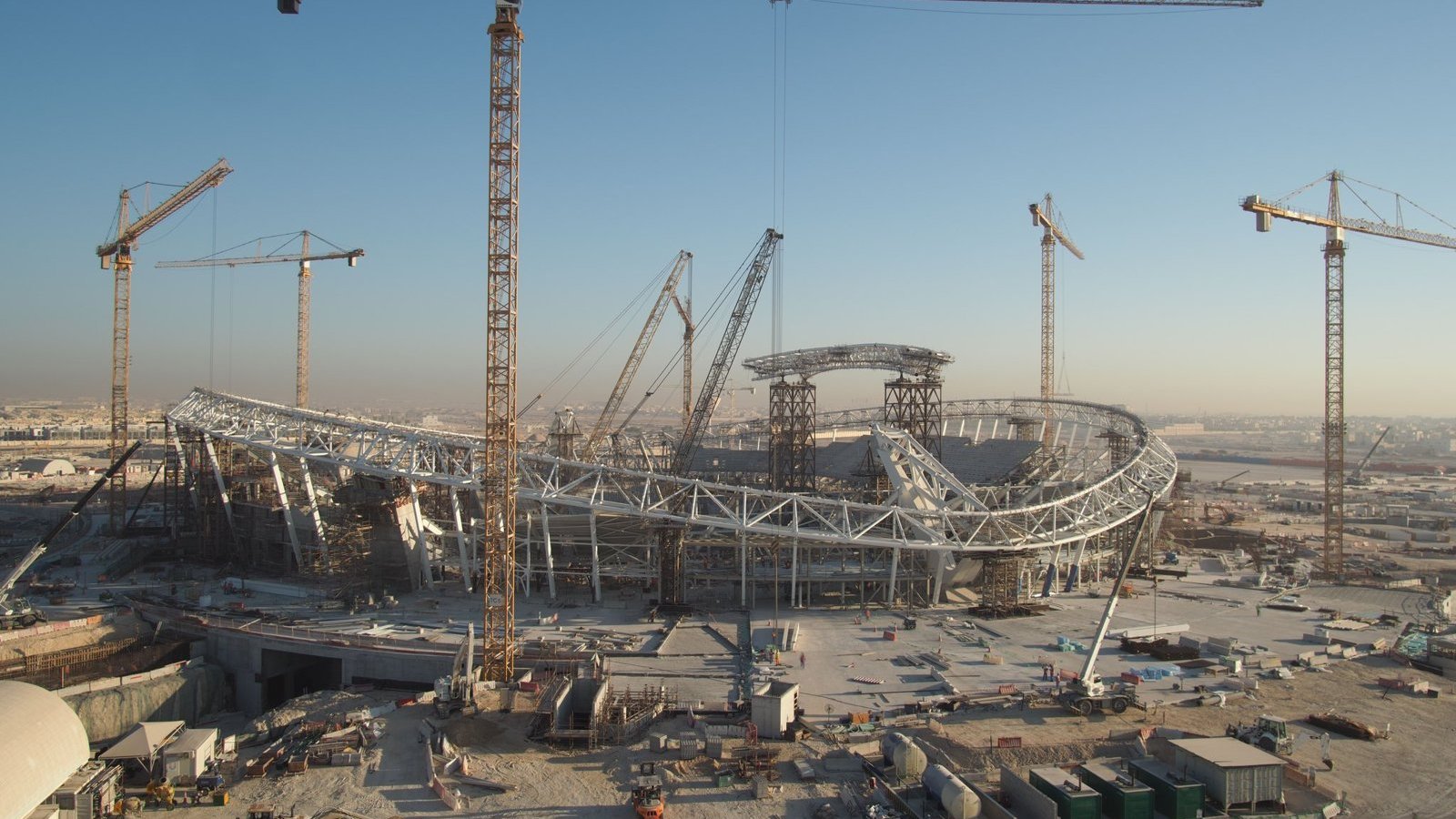 L'image montre un grand chantier de construction d'un stade, avec plusieurs grues en fonctionnement. Le stade a une structure moderne et architecturale, avec des éléments en acier visibles. En arrière-plan, on peut voir un ciel clair, suggérant qu'il fait jour. Le site est entouré de divers matériaux de construction et d'équipements, ce qui indique que le projet est encore en cours.