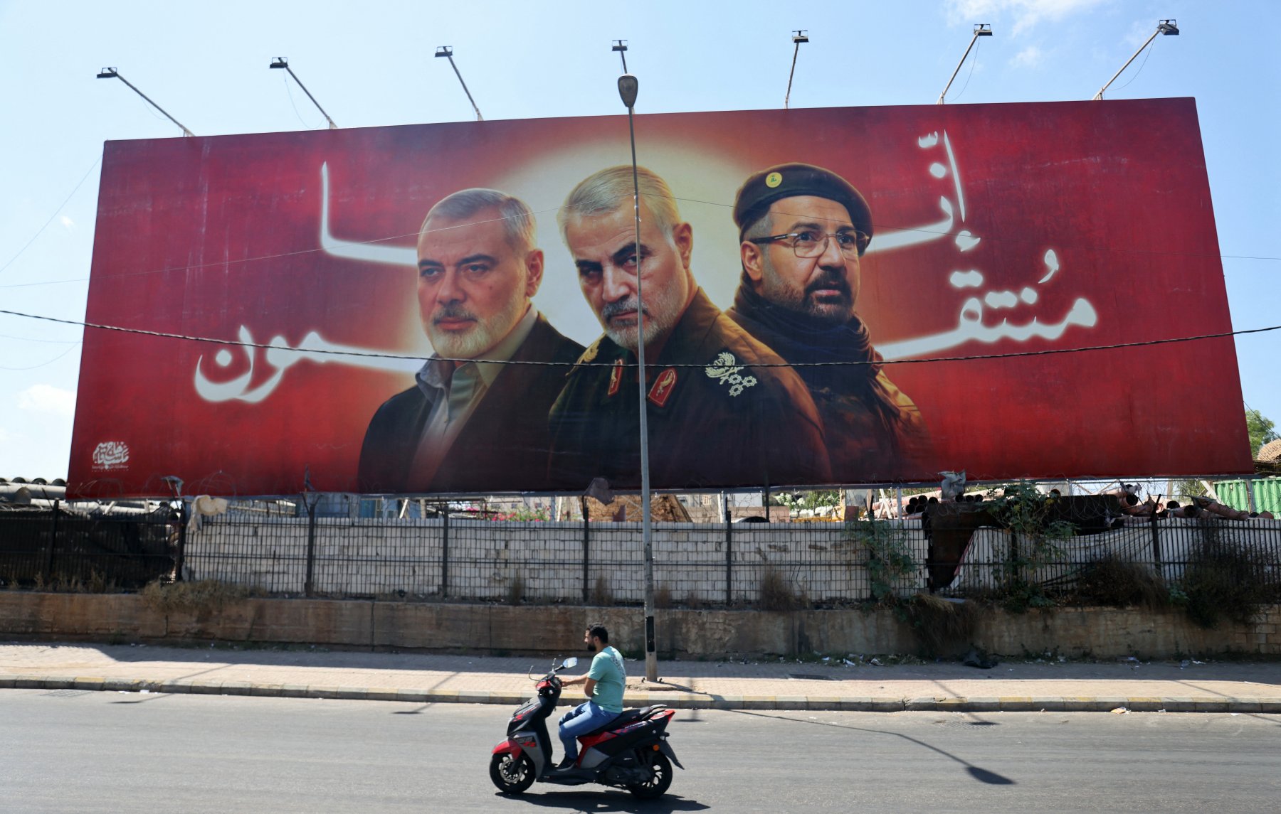 L'image montre une grande affiche publicitaire avec trois portraits d'hommes, probablement des figures militaires ou politiques, sur fond rouge. Un homme à moto passe devant cette affiche, qui semble être placée le long d'une route. L'environnement semble urbain, avec des déchets visibles au fond.