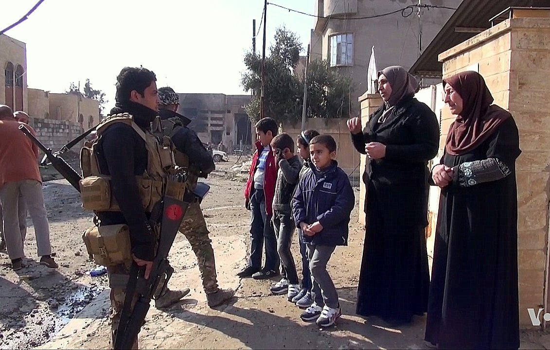 L'image montre une scène dans une rue dévastée, probablement dans une zone de conflit. Un soldat armé est debout, en uniforme militaire, tenant son arme. Il semble interagir avec deux femmes voilées qui lui parlent. À proximité, plusieurs enfants, dont deux garçons, regardent la scène. Le décor est marqué par des bâtiments endommagés et des débris qui témoignent d'une situation difficile. L'atmosphère semble tendue mais il y a aussi une interaction sociale entre les personnes présentes.