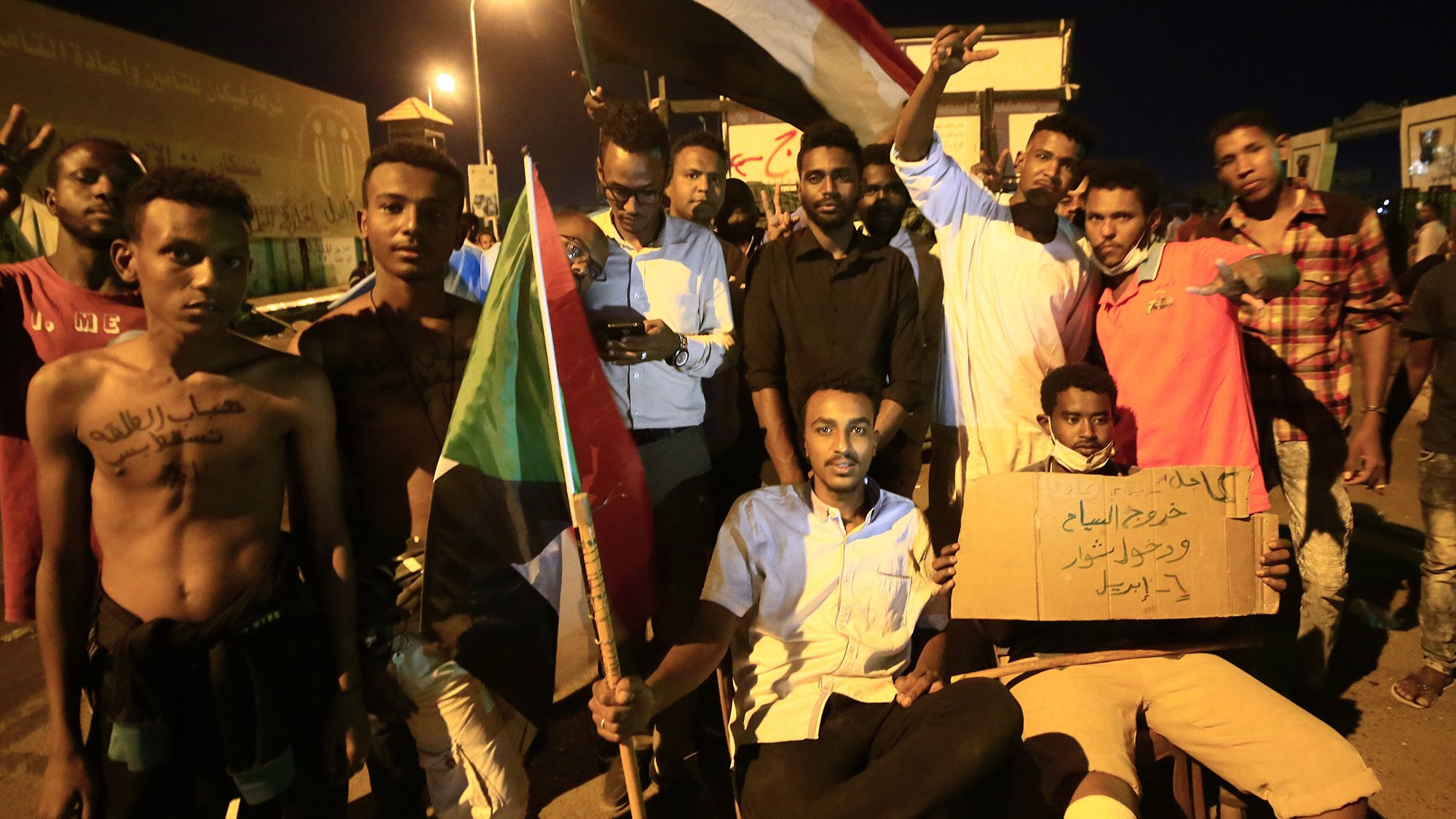 L'image montre un groupe de manifestants soudanais rassemblés lors d'une nuit, tenant des drapeaux et des pancartes. Certains d'entre eux sont debout, affichant des signes de solidarité avec les bras levés, tandis que d'autres sont assis, tenant des panneaux de revendication. L'atmosphère semble énergique et déterminée, incarnant un esprit de protestation et de demande de changement. Les visages des participants expriment diverses émotions, allant de la détermination à l'espoir.