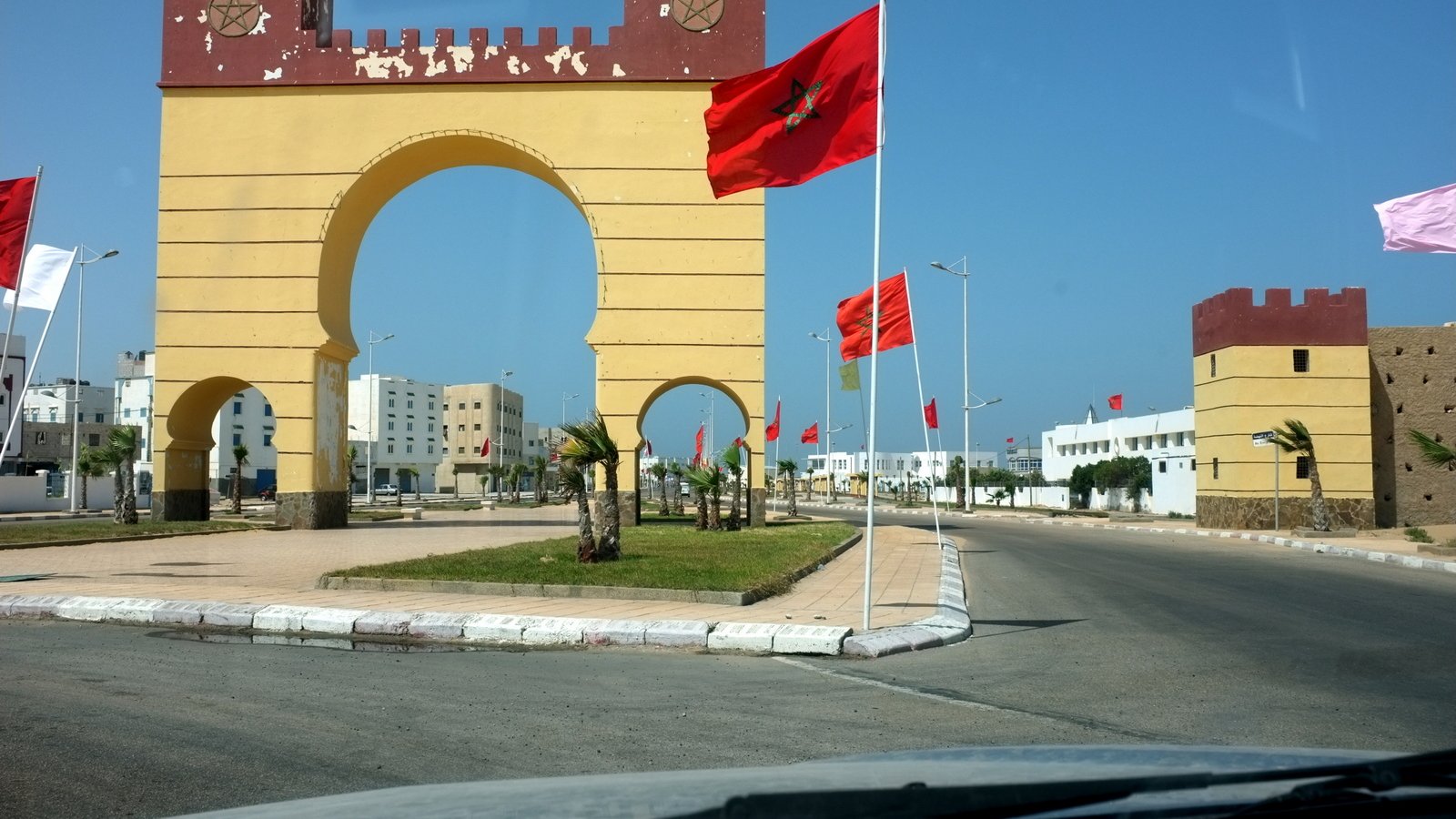 L'image montre un grand arc de triomphe peint en jaune avec des éléments rouges en haut, au centre d'une route. Drapeaux marocains flottent de chaque côté de l'arc, ajoutant une touche nationale à la scène. Des bâtiments modernes peuvent être aperçus au loin, tandis que la route est bordée de palmiers et d'une pelouse bien entretenue. Le ciel est clair et bleu, ce qui donne une ambiance ensoleillée et agréable à l'ensemble.