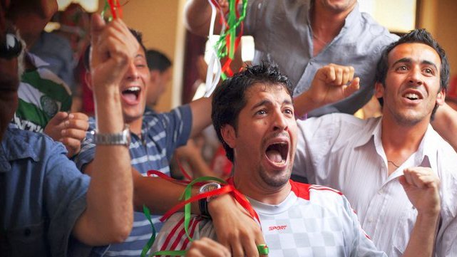 L'image montre un groupe de jeunes hommes dans une ambiance festive. Ils semblent très joyeux et enthousiastes, certains criant et souriant, probablement en train de célébrer un événement sportif. Ils portent des vêtements décontractés et il y a des décorations colorées autour d'eux. Au premier plan, un homme exprime sa joie de manière très expressive, avec les bras levés. En bas, on peut voir un slogan qui dit "LA VIE VOUS APPELLE". L'ensemble de la scène dégage une atmosphère de camaraderie et de bonheur collectif.
