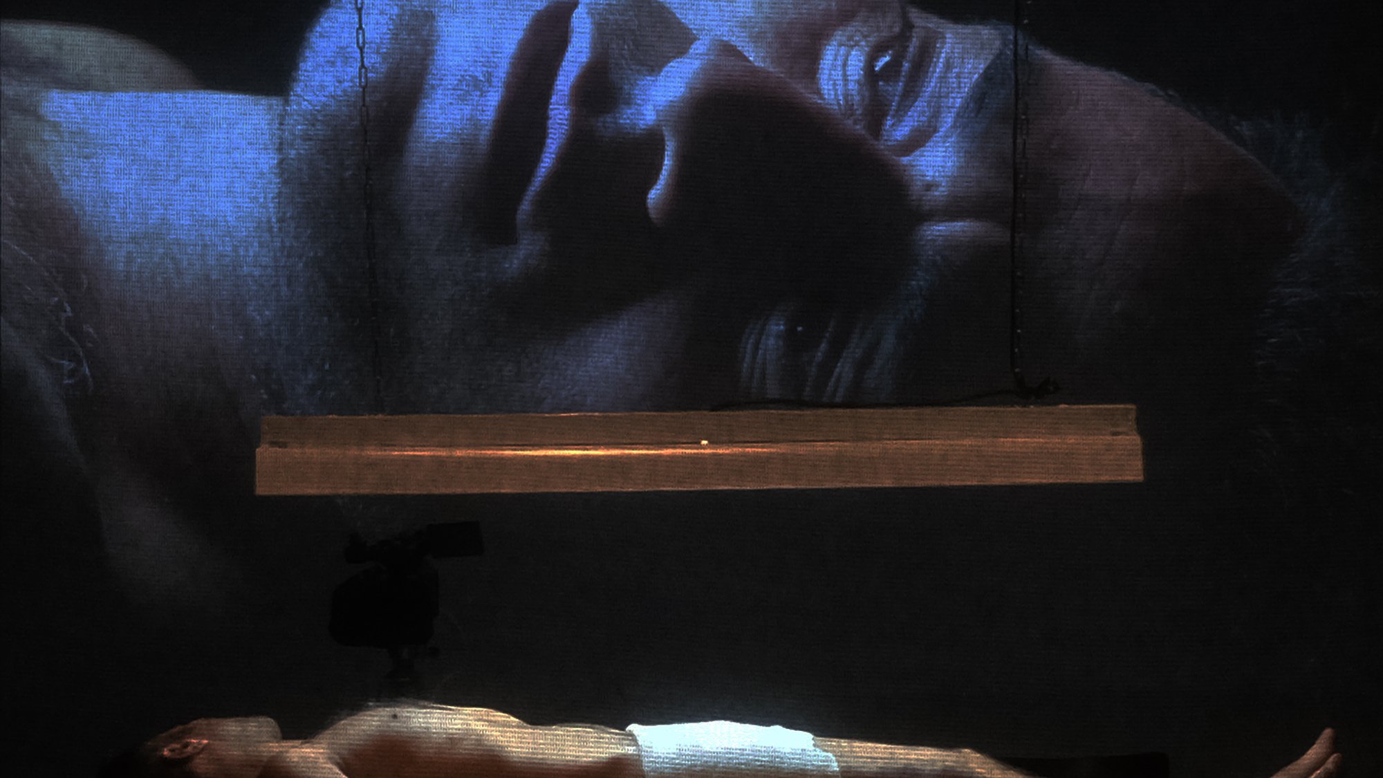 L'image montre un homme allongé sur une surface, partiellement nu, avec une serviette couvrant son corps. Au-dessus de lui, une projection de son visage est affichée sur un grand écran, créant un effet visuel puissant. La lumière provient d'une source suspendue, ajoutant une atmosphère dramatique à la scène. Les détails du visage projeté sont clairement visibles, tandis que l'homme au sol semble dans un état de vulnérabilité. L'ensemble évoque des thèmes de réflexion, d'introspection ou de méditation.