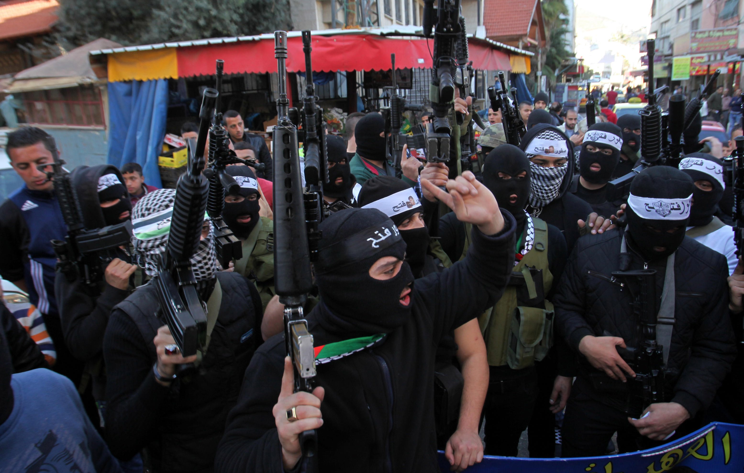 L'image montre un groupe de personnes portant des masques noirs et tenant des armes. Elles se rassemblent dans une rue, avec des bâtiments et des commerçants en arrière-plan. L'atmosphère semble tendue, et leurs expressions indiquent une certaine détermination ou colère. Des drapeaux et des banderoles sont visibles, suggérant un contexte de manifestation ou de rassemblement politique.