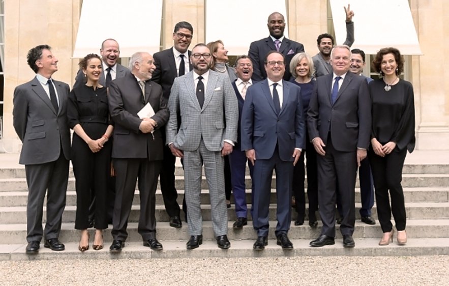 L'image montre un groupe de personnes posant sur des marches devant un bâtiment, probablement lors d'un événement officiel. Au centre, deux hommes se tiennent côte à côte, avec des costumes élégants. Les autres membres du groupe sont habillés de manière formelle ou semi-formelle et sourient pour la photo. L'ambiance semble conviviale, et l'un des hommes à l'arrière fait un signe de paix avec sa main. Les lieux sont lumineux et semblent bien entretenus, ajoutant à l'atmosphère de l'événement.