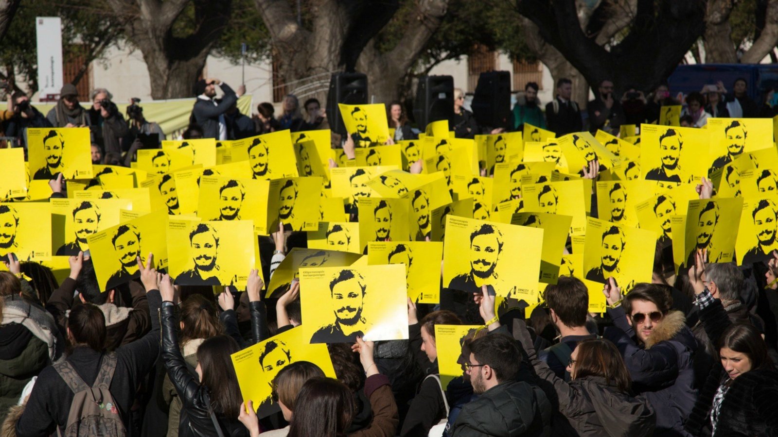 L'image montre une grande foule rassemblée, tenant des pancartes jaunes avec un visage représenté en noir. Les participants semblent mobilisés pour une cause, se tenant la main levée en signe de protestation ou de soutien. En arrière-plan, on peut voir d'autres personnes et des arbres, suggérant que l'événement a lieu en plein air. L'atmosphère paraît engagée et déterminée.