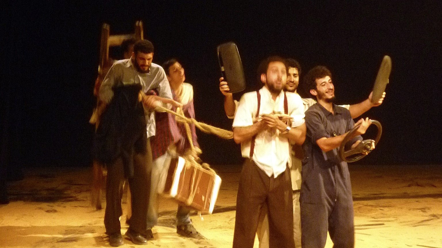 L'image montre un groupe de personnes sur une scène de théâtre, en train de créer une sorte de performance. Ils semblent emporter différents objets, comme des chaises et une valise, en formant une sorte de procession. La scène est éclairée de manière à mettre en évidence leur mouvement, tandis que le fond est sombre, ce qui attire l'attention sur les acteurs. L'ensemble donne une impression dynamique et dramatique, évoquant une histoire ou une interaction entre les personnages.