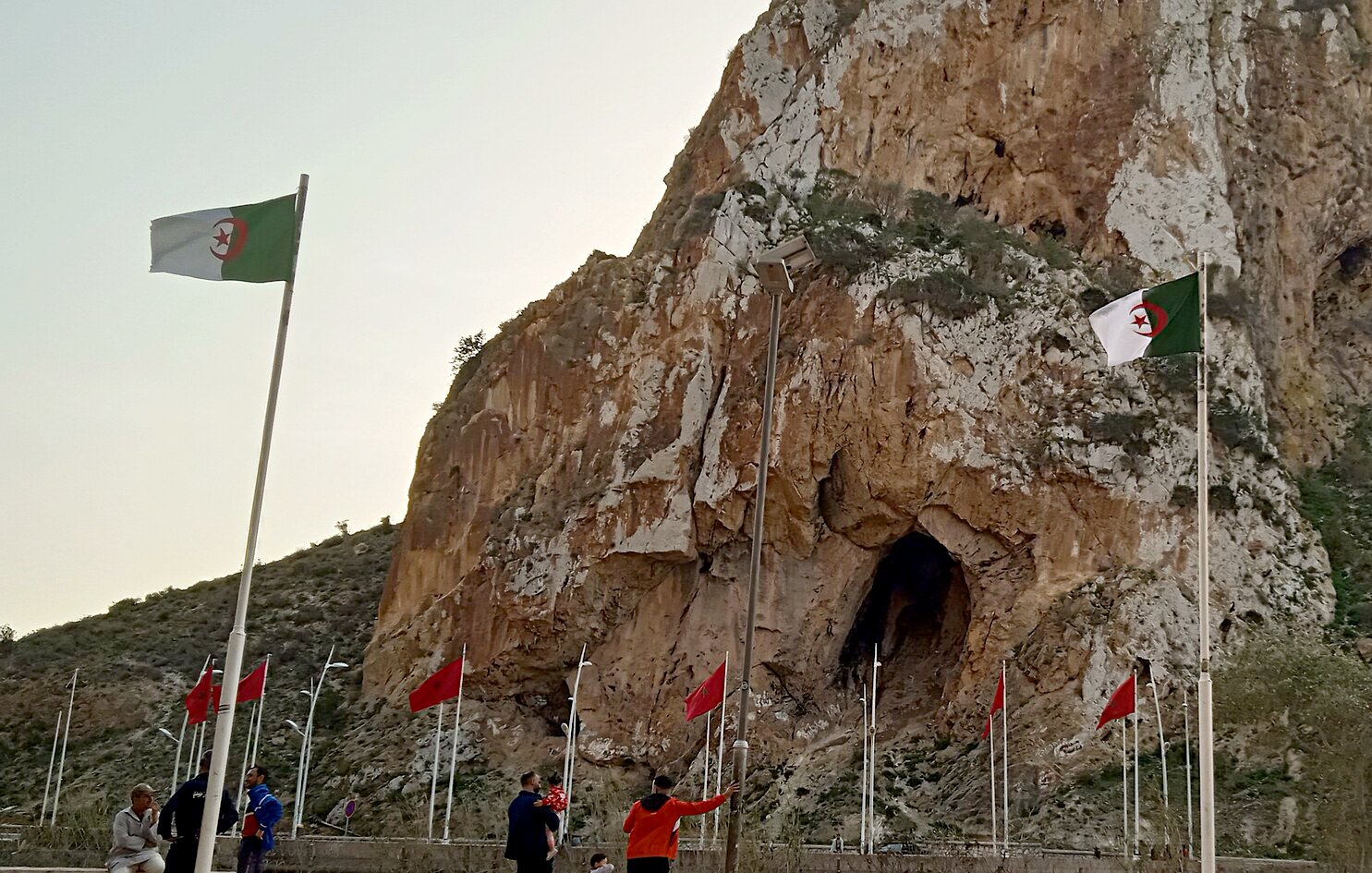L'image montre une imposante falaise avec une grande ouverture, probablement une grotte, qui se trouve à sa base. Des drapeaux algériens flottent autour, ajoutant une touche de couleur au paysage. Au premier plan, on peut voir des personnes marchant près des drapeaux. Le ciel est teinté de lumière douce, suggérant peut-être un lever ou un coucher de soleil. L'ensemble évoque un lieu naturel remarquable avec une signification culturelle ou historique.