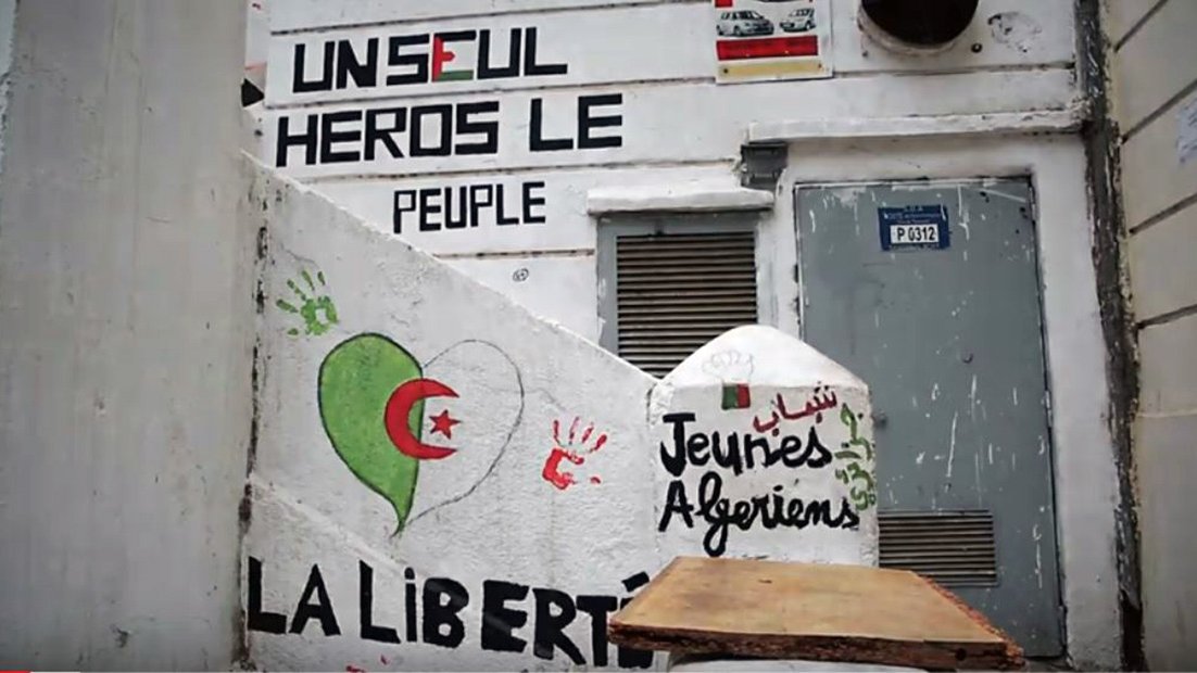 L'image montre un mur peint avec plusieurs graffitis. On y trouve des mots et des phrases exprimant des messages politiques et sociaux. Un dessin représentant un cœur dans les couleurs du drapeau algérien (rouge et vert) est visible. Il y a aussi une inscription en arabe, ainsi que des mots tels que "Jeunies Algériens" et "LA LIBERTÉ". Le fond suggère un lieu qui pourrait être un espace public, témoignant d'une expression artistique et militante.