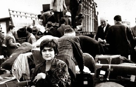 L'image montre une scène de personnes en situation de déplacement. Au premier plan, une jeune fille tient un animal, probablement un chat, dans ses bras, et elle est entourée de nombreux bagages, valises et paquets. En arrière-plan, on aperçoit d'autres personnes, certains sont en train de charger ou de décharger des objets d'un véhicule. L'atmosphère est chargée, et l'image semble capturer un moment de transition ou d'exode, évoquant des sentiments de nostalgie et de désespoir. Les tonalités en noir et blanc ajoutent une dimension dramatique à la scène.