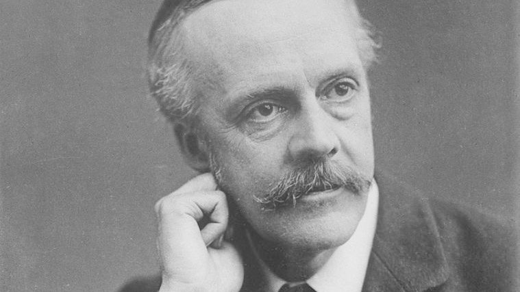 L'image montre un homme en costume vêtu d'une veste sombre et d'une cravate. Il a des moustaches bien entretenues et les cheveux légèrement grisonnants. Il pose en penchant légèrement la tête sur la main, affichant une expression pensive. Le fond est neutre, mettant en valeur son visage et son regard pensif.