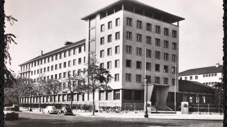 L'image représente un bâtiment architectural de plusieurs étages, probablement de style moderne. Il est photographié sous une lumière naturelle, mettant en avant sa façade aux lignes géométriques. Le bâtiment semble être entouré d'arbres et de quelques véhicules anciens, ce qui donne une ambiance d'époque. On peut également apercevoir des éléments de la rue, comme des lampadaires et éventuellement d'autres bâtiments en arrière-plan. L'ensemble de l'image présente une esthétique monochrome, accentuant le caractère historique de la scène.