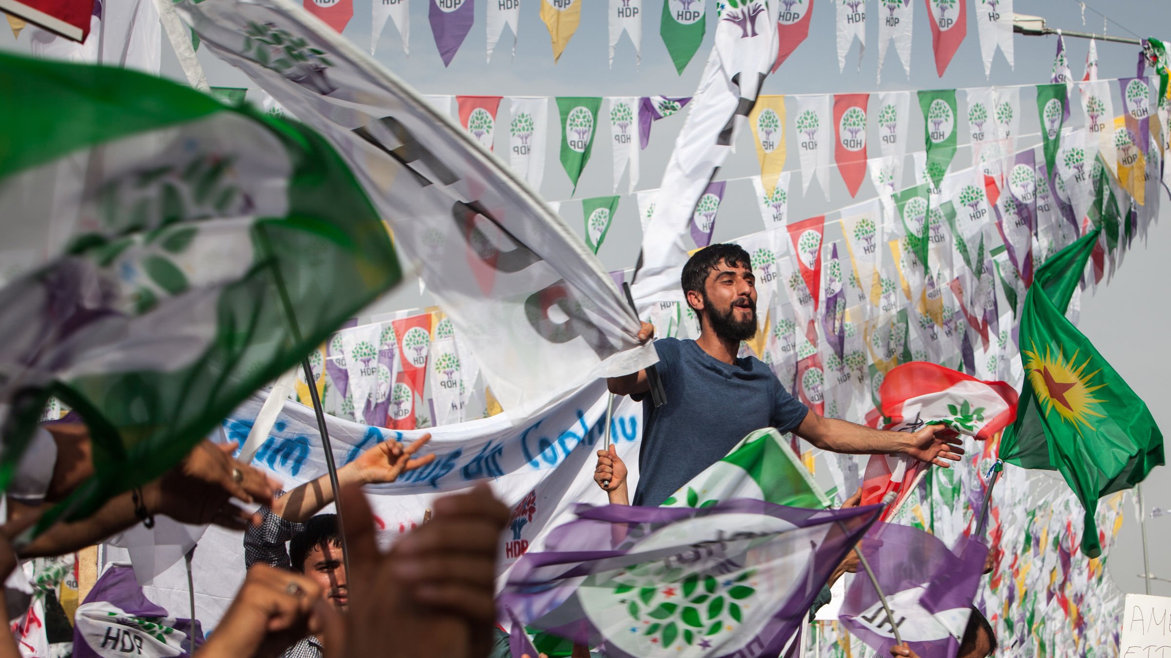 L'image montre une foule rassemblée lors d'un événement politique ou d'une manifestation. On peut voir des personnes brandissant des drapeaux, dont certains portent les couleurs et les symboles associés à un parti. L'ambiance semble festive et engagée, avec des individus visibles qui expriment leur enthousiasme, certains levant les bras ou chantant. En arrière-plan, des fanions colorés flottent, ajoutant à l'atmosphère vibrante de l'événement.