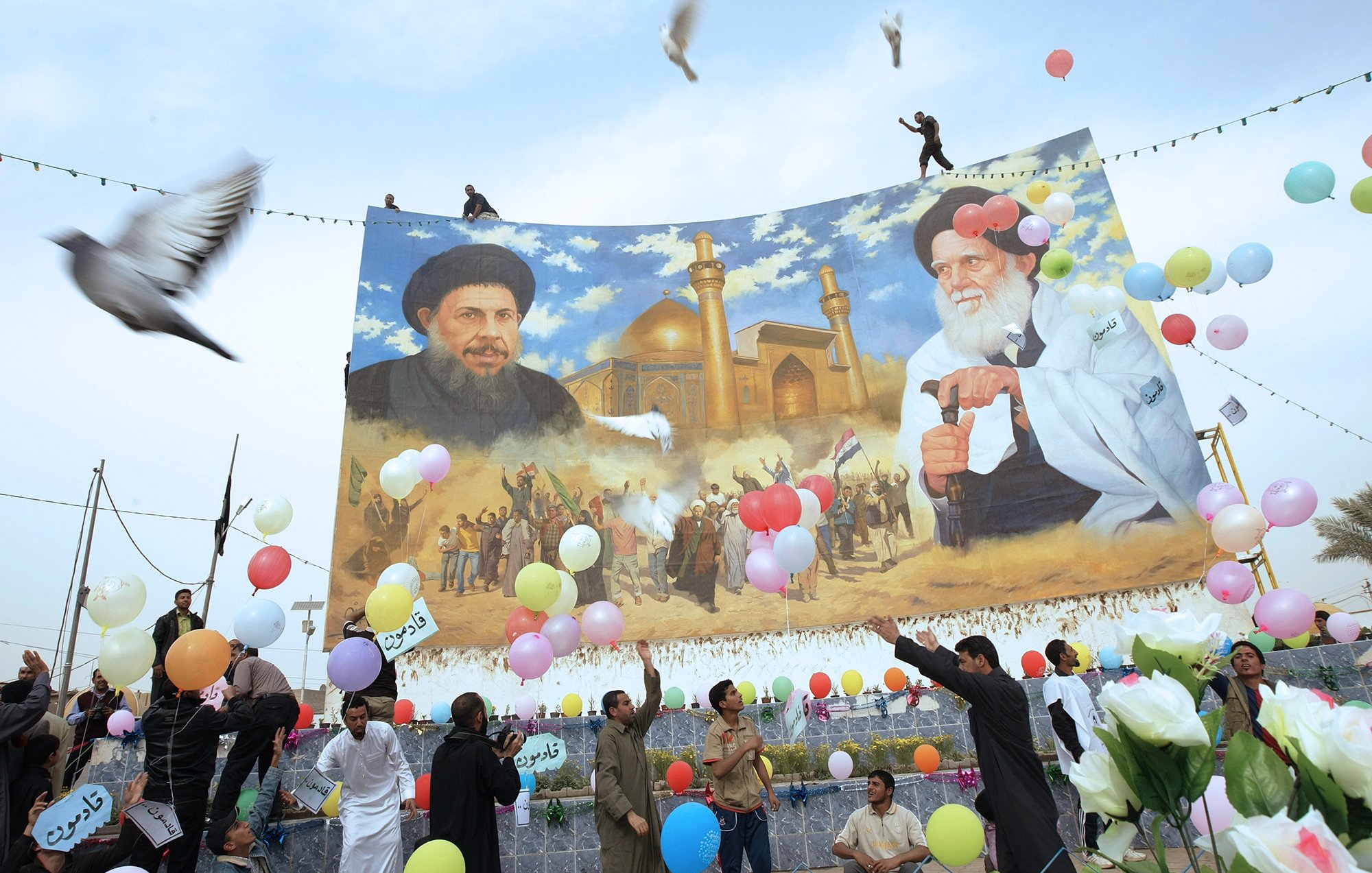 L'image montre une scène festive en extérieur avec un grand panneau représentant deux figures, probablement des leaders ou des personnalités importantes. Au-dessous de ce panneau, des gens célèbrent, tenant des ballons colorés et des fleurs. Certaines personnes semblent danser ou exprimer leur joie. En arrière-plan, il y a un décor architectural qui semble être un bâtiment religieux, avec des minarets visibles. Des colombes volent également dans le ciel, ajoutant une dimension symbolique à l'événement.