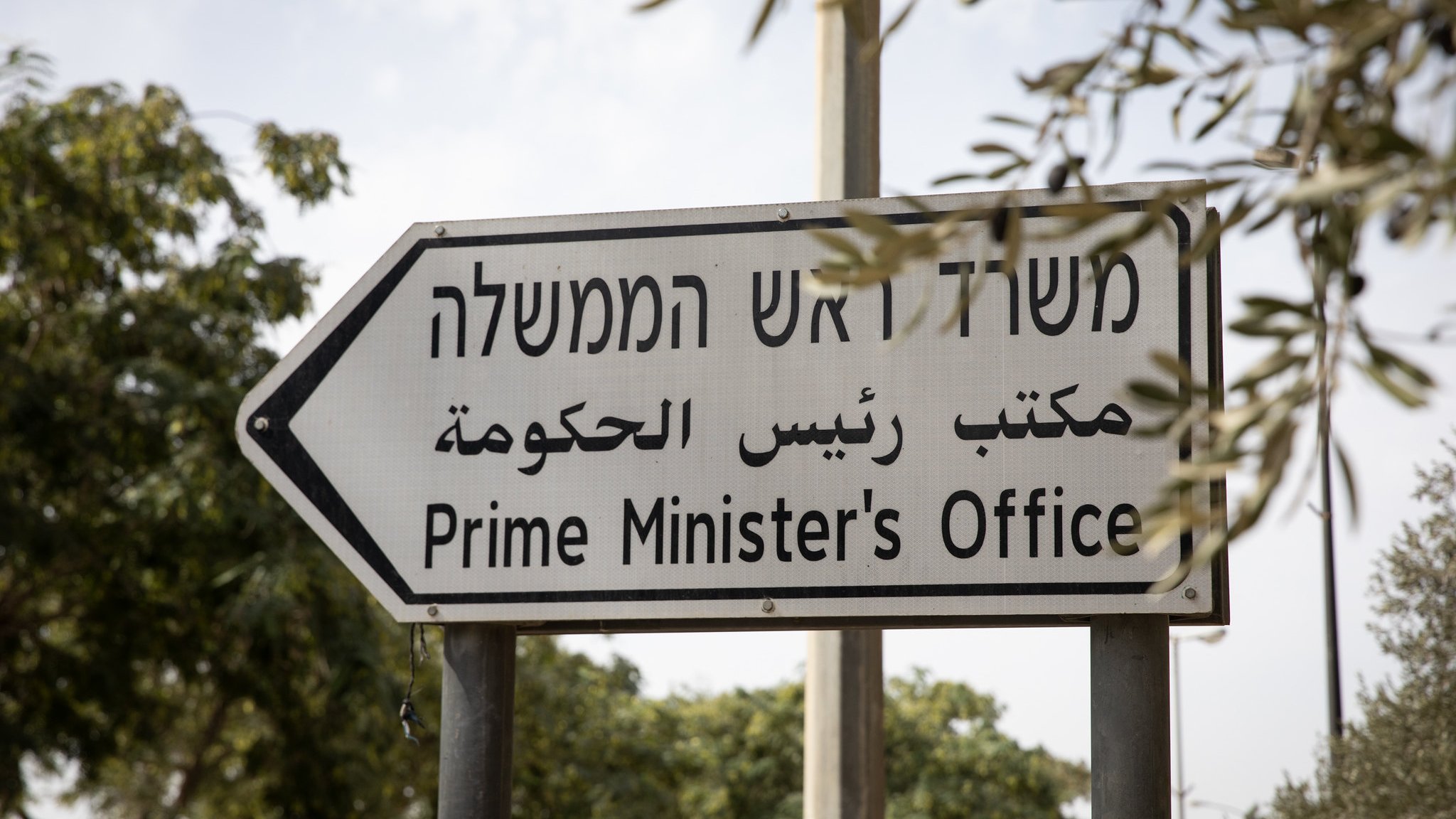 L'image montre un panneau de signalisation indiquant le "Bureau du Premier Ministre". Ce panneau est trilingue, avec des inscriptions en hébreu, en arabe et en anglais, ce qui reflète la diversité linguistique de la région. Le panneau est installé dans un environnement avec des arbres en arrière-plan.