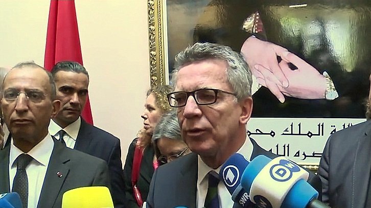 L'image montre un groupe de personnes lors d'une conférence de presse ou d'un événement officiel. Au premier plan, un homme s'exprime devant des caméras et des micros, entouré de plusieurs autres individus qui semblent attentifs. En arrière-plan, on peut voir un portrait ou une image représentant des mains avec des symboles significatifs, ainsi qu'un drapeau. L'atmosphère semble formelle, probablement liée à des discussions d'ordre politique ou diplomatique.