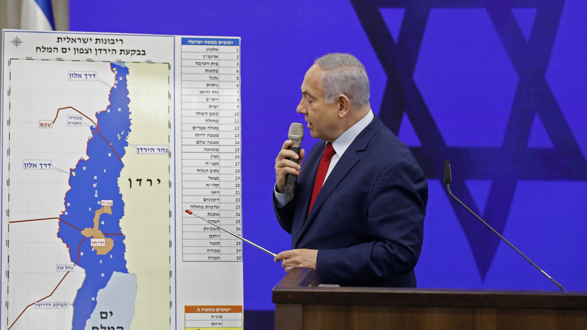 L'image montre un homme debout devant un tableau graphique. Il semble s'adresser à un auditoire, tenant un pointeur dans sa main. Le tableau derrière lui illustre une carte, probablement liée à une question de territoire, avec des annotations en hébreu. On aperçoit également un symbole du judaïsme en arrière-plan. L'homme porte une cravate rouge et un costume sombre.