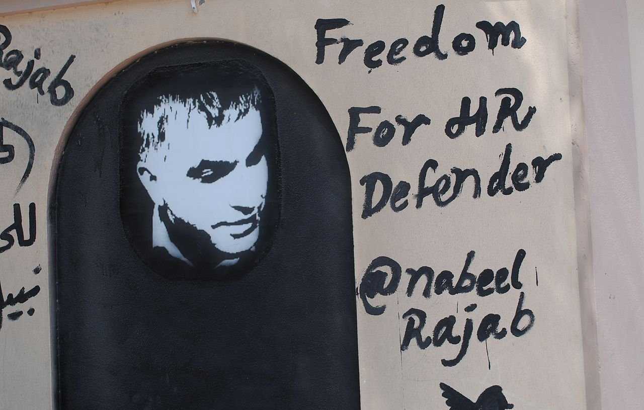 L'image représente un graffiti sur un mur. On y voit un portrait en noir et blanc d'un homme, accompagné d'un texte écrit en anglais. Le message indique "Freedom For HR Defender" (Liberté pour le défenseur des droits de l'homme) et mentionne un nom d'utilisateur Twitter "@nabeelrajab". Le style artistique est simple, avec un fond sombre et des lettres noires qui ressortent sur la surface claire du mur. Cela semble être une œuvre engagée, soulignant le soutien à la défense des droits humains.