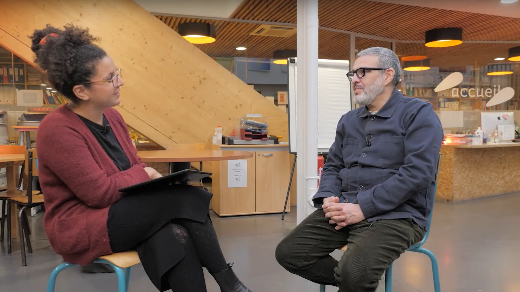 L'image montre deux personnes assises dans un espace intérieur. À gauche, une femme avec des cheveux bouclés et des lunettes, vêtue d'un pull rouge, tient une tablette. À droite, un homme portant des lunettes et une chemise bleu foncé est assis, les mains sur ses genoux. L'environnement est moderne, avec des murs en bois et des éléments de décoration qui donnent une ambiance chaleureuse. Cela semble être une interview ou une discussion.