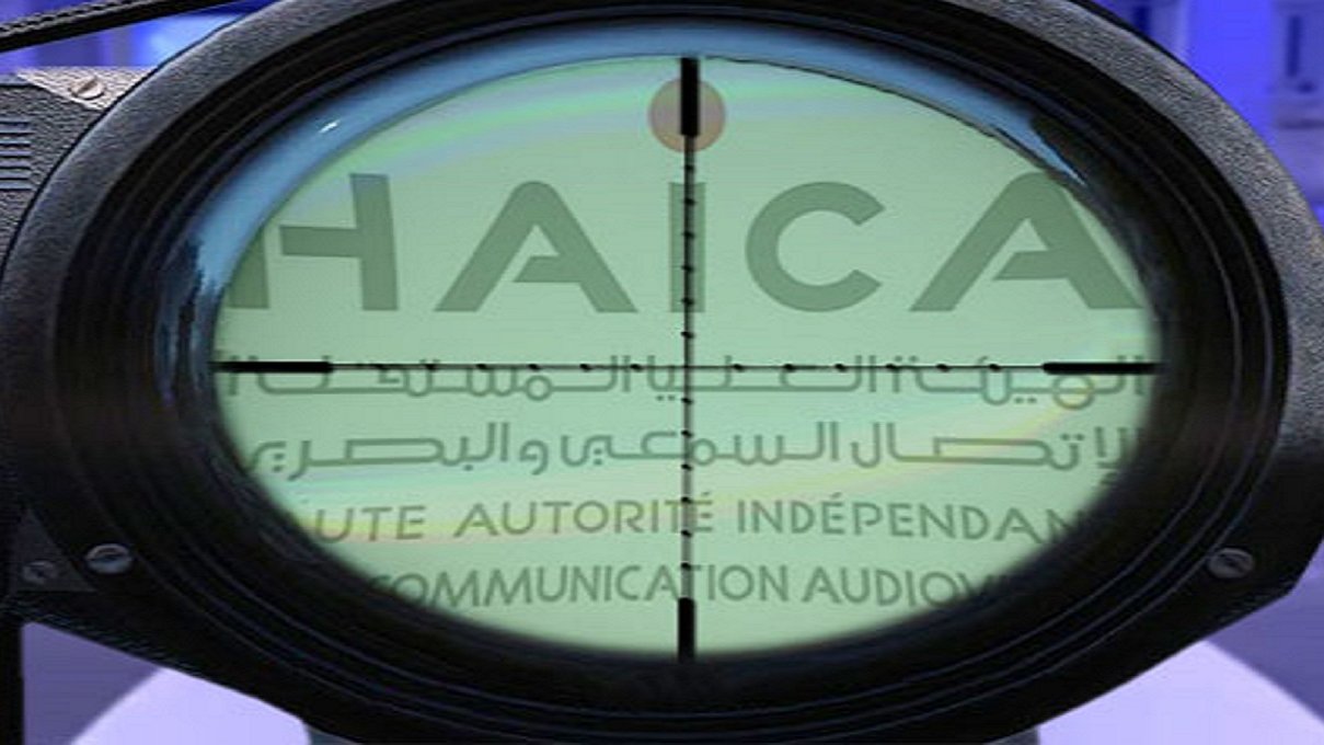 L'image montre un réticule de visée, typique d'une lunette de tir, qui se superpose à un fond avec le mot "HAICA" visible. Ce mot est associé à une autorité indépendante en matière de communication auditive, peut-être en rapport avec la régulation des médias. Le contraste entre l'optique et le texte suggère un thème de surveillance ou de contrôle. Le fond semble comporter divers éléments, peut-être liés à des événements ou des institutions médiatiques.