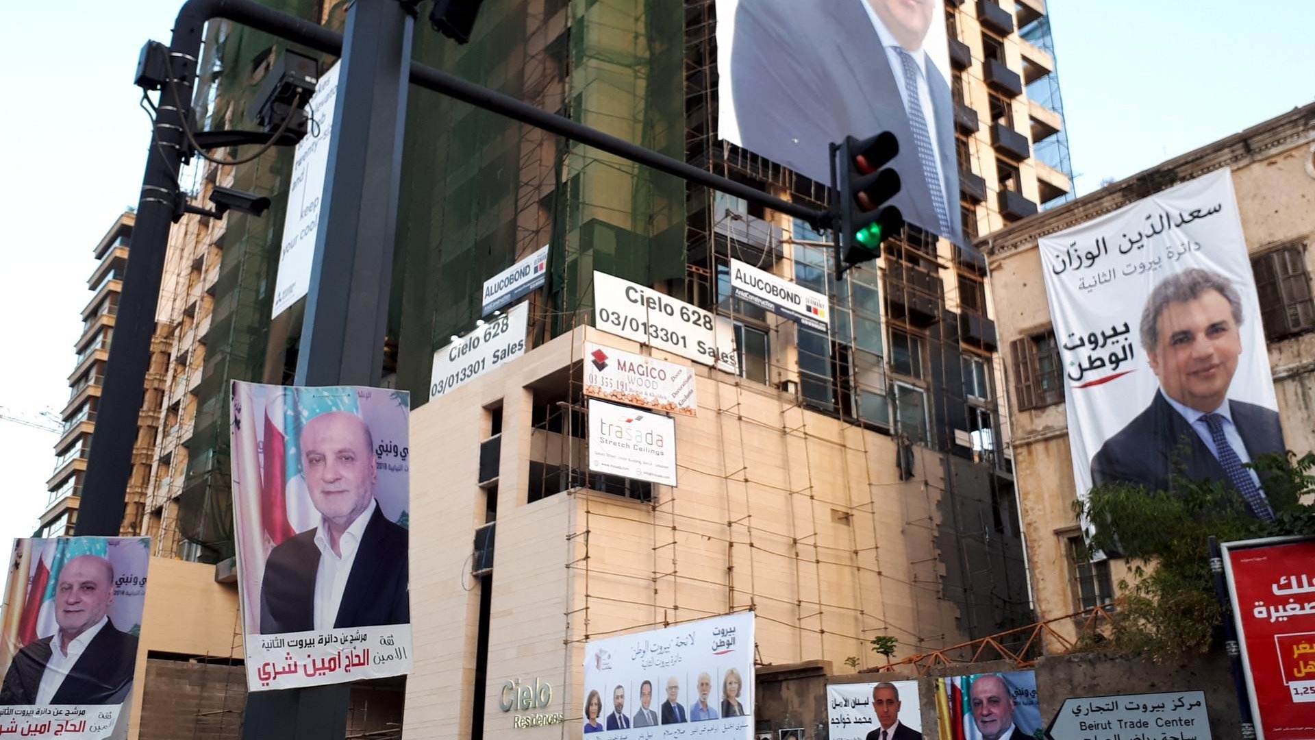 L'image montre un immeuble en construction ou rénové, arborant plusieurs affiches et panneaux publicitaires. Les affiches présentent des portraits de différentes personnes, probablement des candidats ou des figures politiques, accompagnés de textes en arabe. La scène donne l'impression d'une campagne électorale ou d'une promotion politique. Il y a également un feu de signalisation visible à gauche, ce qui indique que cette image est prise dans un environnement urbain.