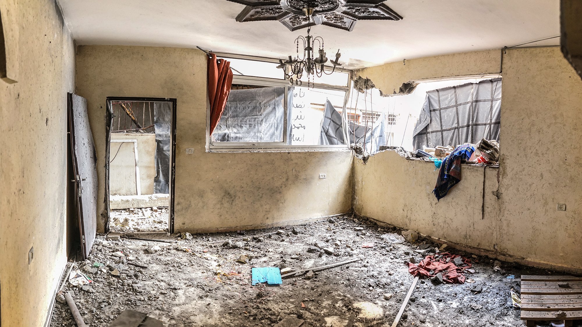 L'image montre une pièce intérieure en mauvais état. Les murs sont abîmés et la peinture est écaillée. Il y a des débris et des gravats sur le sol, ce qui indique un manque d'entretien ou des dommages récents. Une fenêtre est brisée, laissant passer la lumière et révélant des rideaux déchirés. Des vêtements et d'autres objets sont éparpillés, renforçant l'impression de désordre et d'abandon. Un lustre pend du plafond, ajoutant un contraste avec l'état délabré de l'espace. L'ambiance générale semble triste et négligée.