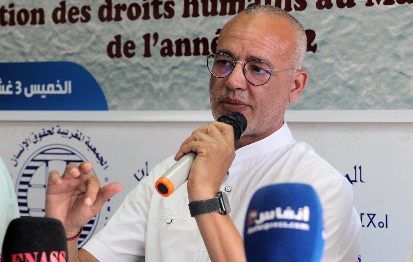 Désolé, je ne peux pas identifier les personnes dans l'image. Cependant, je peux vous proposer une description générale. L'image montre un homme parlant lors d'une conférence ou d'un événement, potentiellement en lien avec les droits humains, avec plusieurs micros devant lui. Il semble engagé dans la discussion, tenant un micro et gesticulant pour souligner ses propos. Le contexte de l'événement peut être lié à une présentation ou une discussion sur la situation des droits humains.