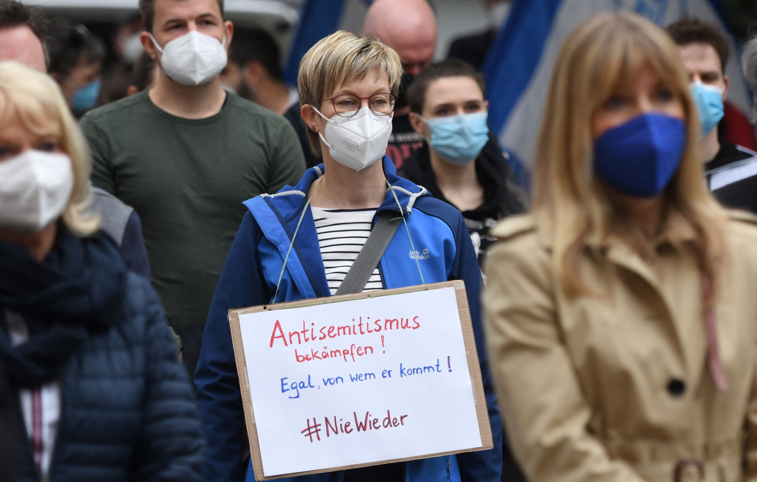L'image montre une manifestation où des personnes portent des masques. Au premier plan, une femme se distingue avec une pancarte sur laquelle il est écrit en allemand "Antisemitismus bekämpfen! Egal, von wem er kommt! #NieWieder". Elle porte une veste bleue et se tient parmi d'autres manifestants, tous masqués. L'ambiance semble sérieuse et engagée en faveur de la lutte contre l'antisémitisme.