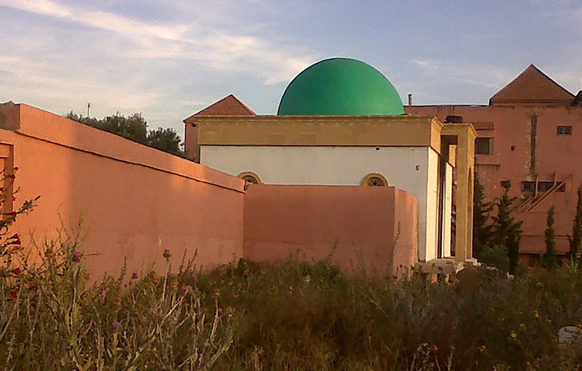 L'image montre un bâtiment avec une coupole verte, partiellement cachée par un mur. Le bâtiment semble être de style architectural traditionnel, avec des murs peints en rose et des fenêtres rondes. On remarque également une végétation sauvage devant, ajoutant un aspect naturel à la scène. En arrière-plan, on aperçoit d'autres structures, suggérant un environnement urbain ou semi-urbain. Le ciel est clair, indiquant une atmosphère paisible.