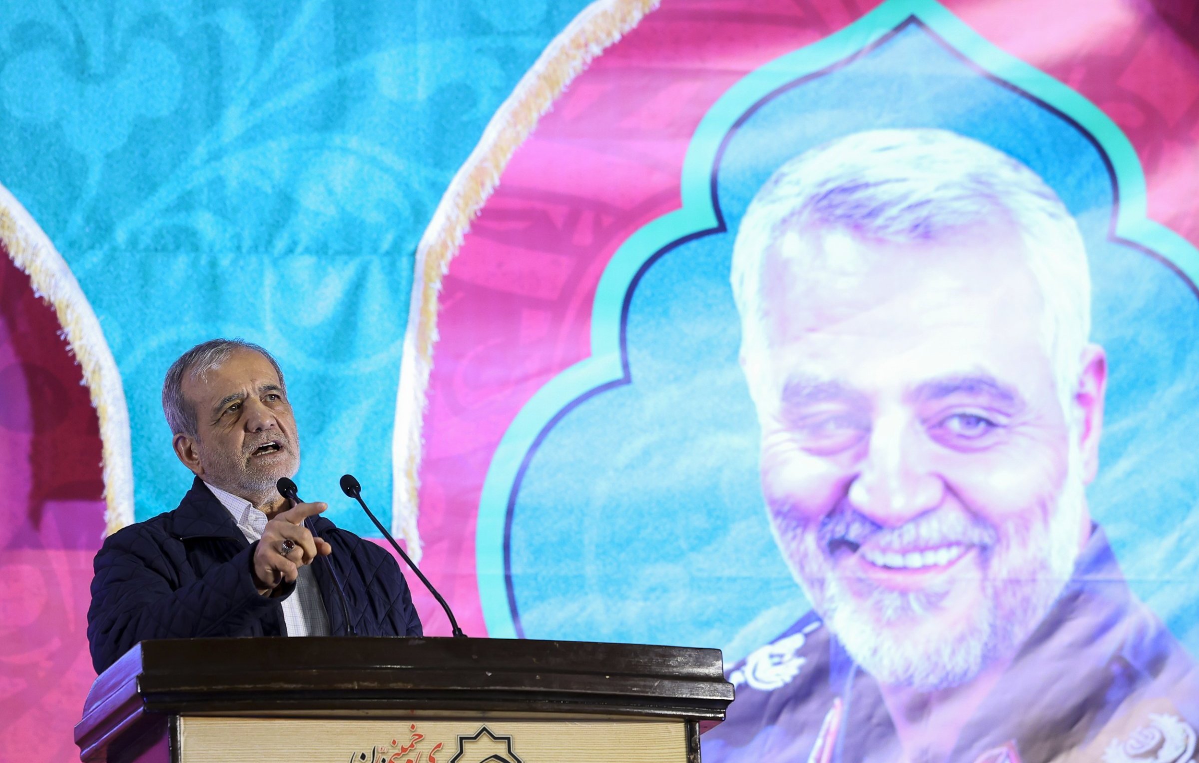 L'image montre un homme en train de s'exprimer lors d'un discours devant un microphone. En arrière-plan, il y a un grand panneau coloré affichant le portrait d'un homme souriant, probablement une figure importante. Le fond est décoré avec des motifs vibrants. L'homme au premier plan semble prendre la parole avec passion, pointant du doigt, ce qui indique qu'il parle probablement d'un sujet important ou émotionnel.