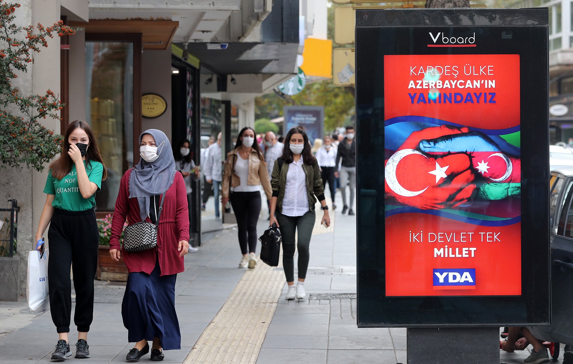 L'image montre une scène de rue animée où des personnes marchent sur un trottoir. À gauche, deux femmes avancent, l'une portant un hijab gris et l'autre en tenue décontractée. À droite, un panneau d'affichage numérique affiche un message en turc, avec des couleurs vives et des symboles représentant les drapeaux de la Turquie et de l'Azerbaïdjan. Le texte semble faire référence à un soutien entre les deux pays. Plusieurs passants peuvent être aperçus en arrière-plan, portant tous des masques de protection.