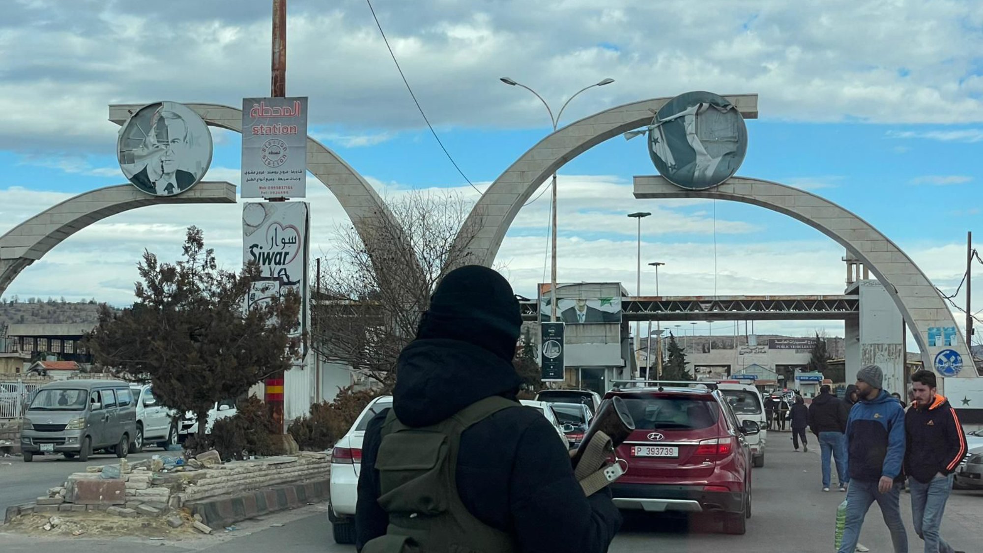 L'image montre un grand arc de triomphe à l'entrée d'une ville, avec des réparations visibles. Deux panneaux ronds sont accrochés en haut, affichant des illustrations. La scène est animée avec des voitures et des passants, sous un ciel nuageux.