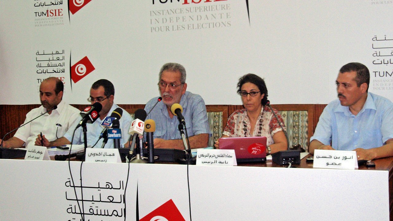 L'image montre une conférence de presse avec plusieurs personnes assises à une table. Un homme au centre semble parler, tandis que les autres personnes à ses côtés écoutent ou prennent des notes. Il y a des microphones sur la table et un arrière-plan avec des logos et du texte en arabe et en français, probablement lié à une institution électorale. L'environnement semble formel et consacré aux affaires publiques.
