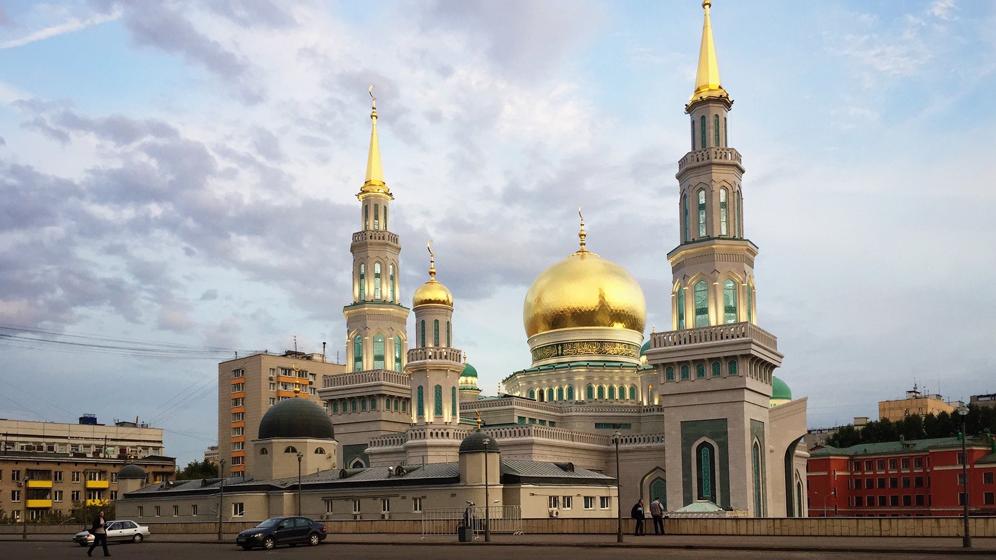 L'image montre une mosquée impressionnante avec des coupoles dorées et plusieurs minarets. Les structures sont ornées de détails architecturaux colorés, et le ciel en arrière-plan est légèrement nuageux, créant une ambiance tranquille. La mosquée se dresse majestueusement en milieu de ville, entourée d'immeubles modernes.