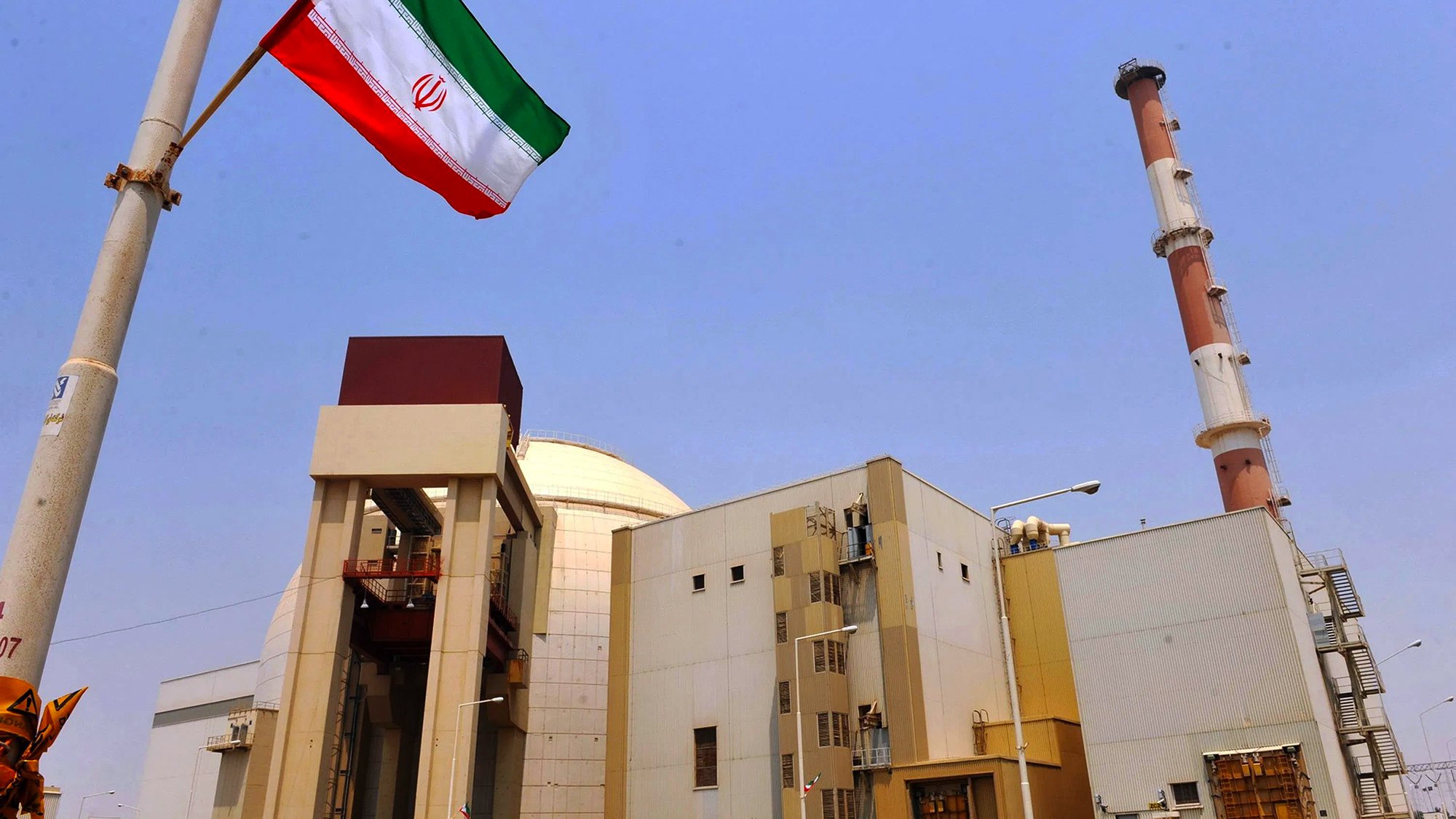 L'image montre une installation industrielle avec des structures modernes. On peut voir une grande cheminée au fond et un bâtiment principal qui semble être lié à l'énergie. Au premier plan, il y a un mât avec le drapeau iranien flottant, qui arbore des bandes rouges, blanches et vertes. Le ciel est dégagé, créant un contraste avec les bâtiments. L'ensemble évoque une infrastructure liée à la production d'énergie, probablement nucléaire.