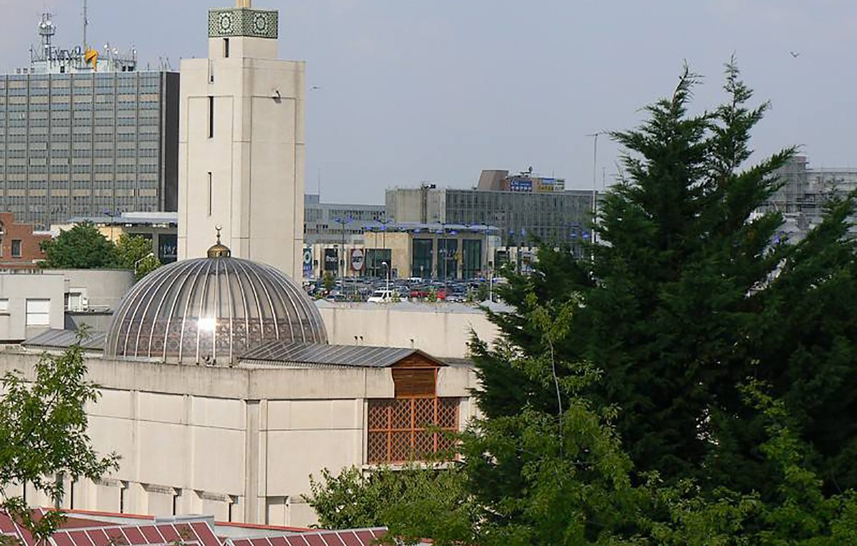 L'image montre un bâtiment architectural avec un dôme et une tour horloge, entouré d'arbres et d'autres structures urbaines en arrière-plan. On peut voir un environnement mixte, combinant des éléments d'architecture moderne et traditionnelle, avec des stations de transports et des bâtiments de bureaux. La scène est bien éclairée, suggérant une journée ensoleillée.