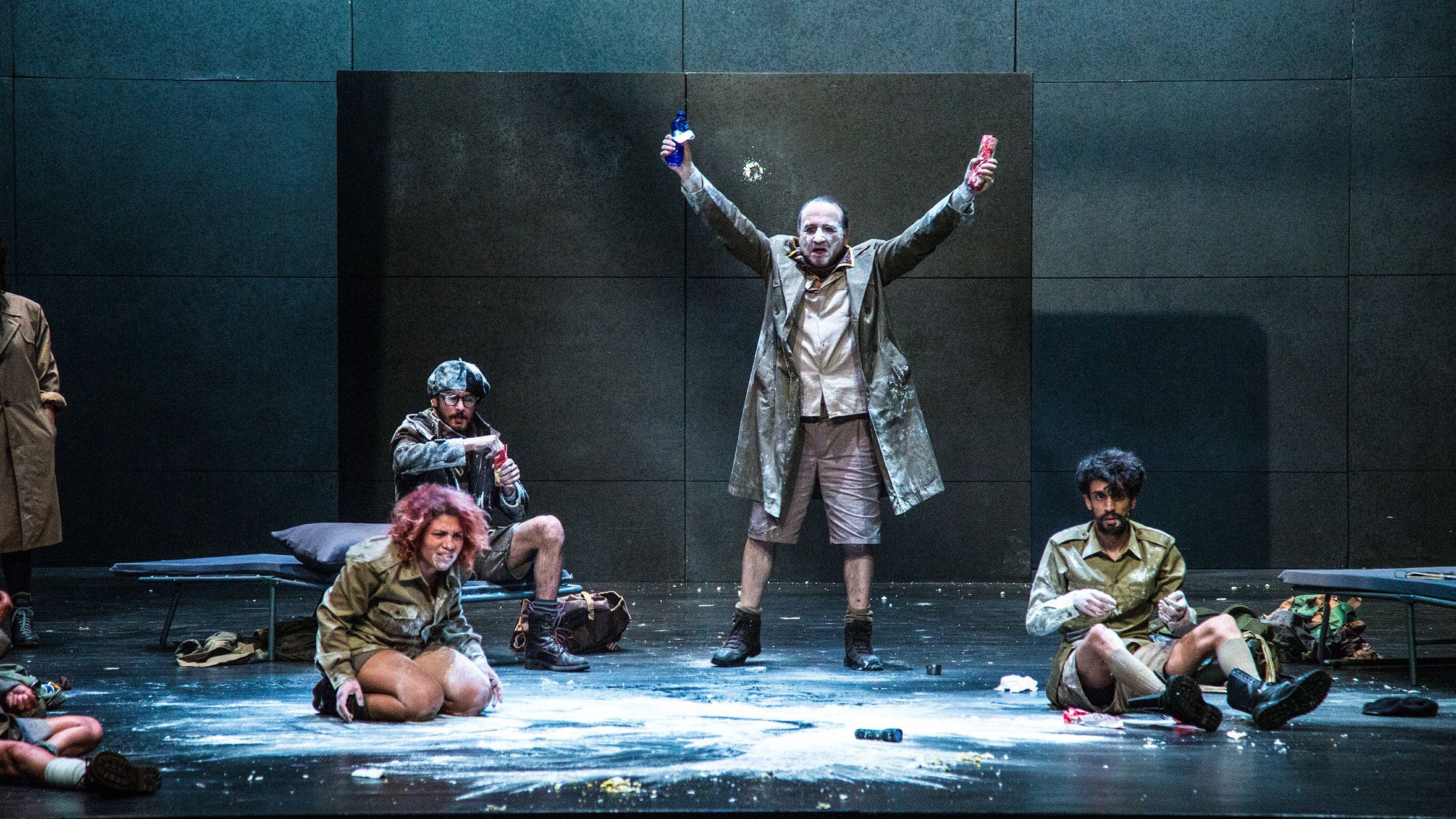 L'image montre une scène de théâtre sur une scène dépouillée. Au centre, un homme en manteau se tient avec les bras levés, tenant une bouteille dans chaque main, ce qui suggère une attitude triomphante ou exubérante. Autour de lui, plusieurs autres personnages sont assis ou accroupis, vêtus de costumes similaires. L'un d'eux semble concentré sur une activité au sol. La scène peut évoquer une ambiance dramatique ou absurde, avec des éléments de désordre comme de la poudre dispersée sur le sol. Les expressions des personnages varient, ajoutant à la dynamique de l'ensemble.