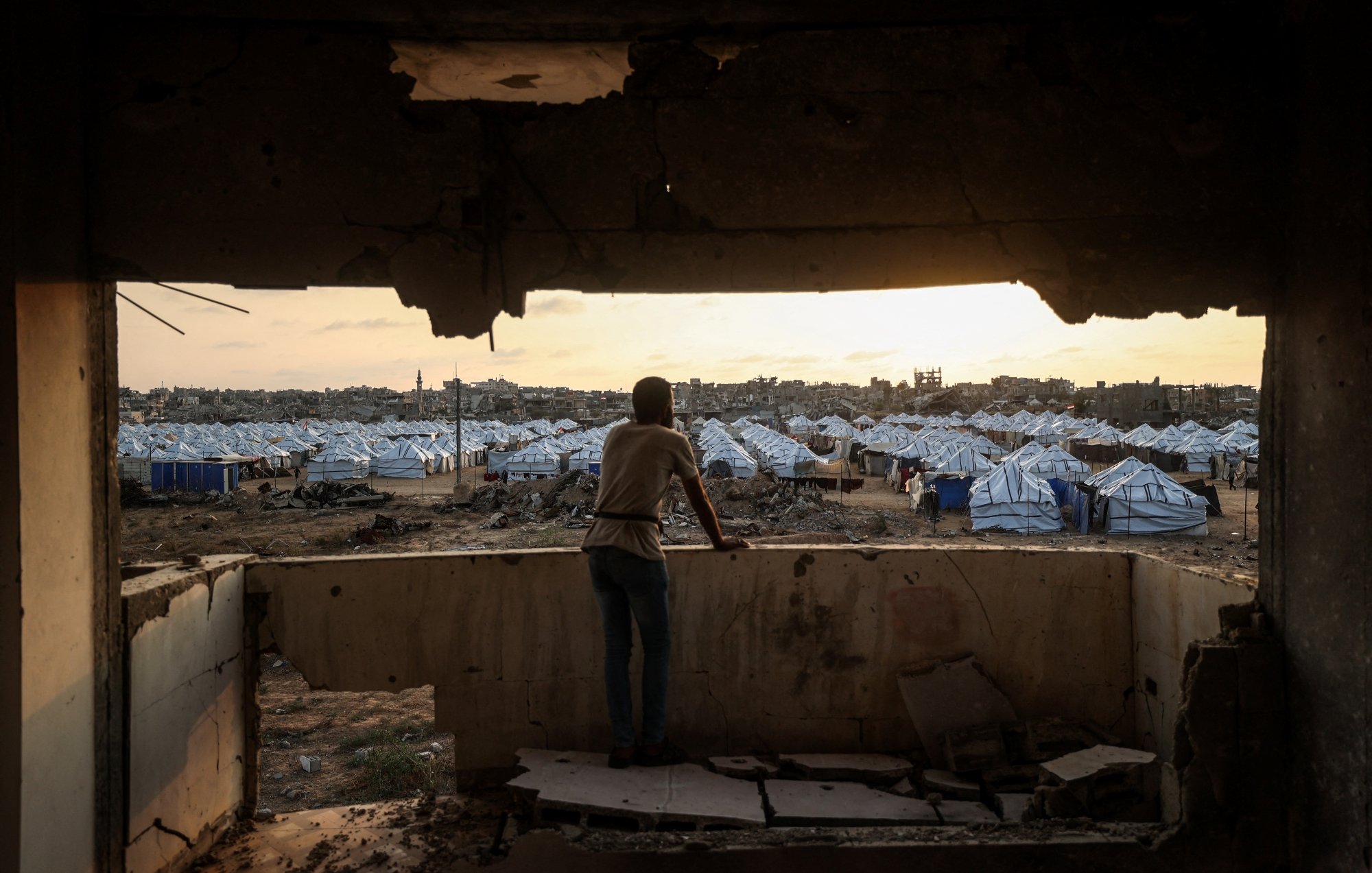 Un jeune observe un camp de tentes depuis une fenêtre endommagée au crépuscule.