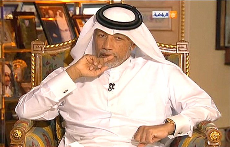 L'image montre un homme assis sur un fauteuil, habillé en costume traditionnel arabe, avec un headdress. Il a une expression pensive et semble concentré, en train de penser ou de réfléchir. En arrière-plan, on peut voir des photographies accrochées au mur, ajoutant une touche personnelle à l'environnement. L'atmosphère semble être celle d'une interview ou d'une discussion sérieuse.