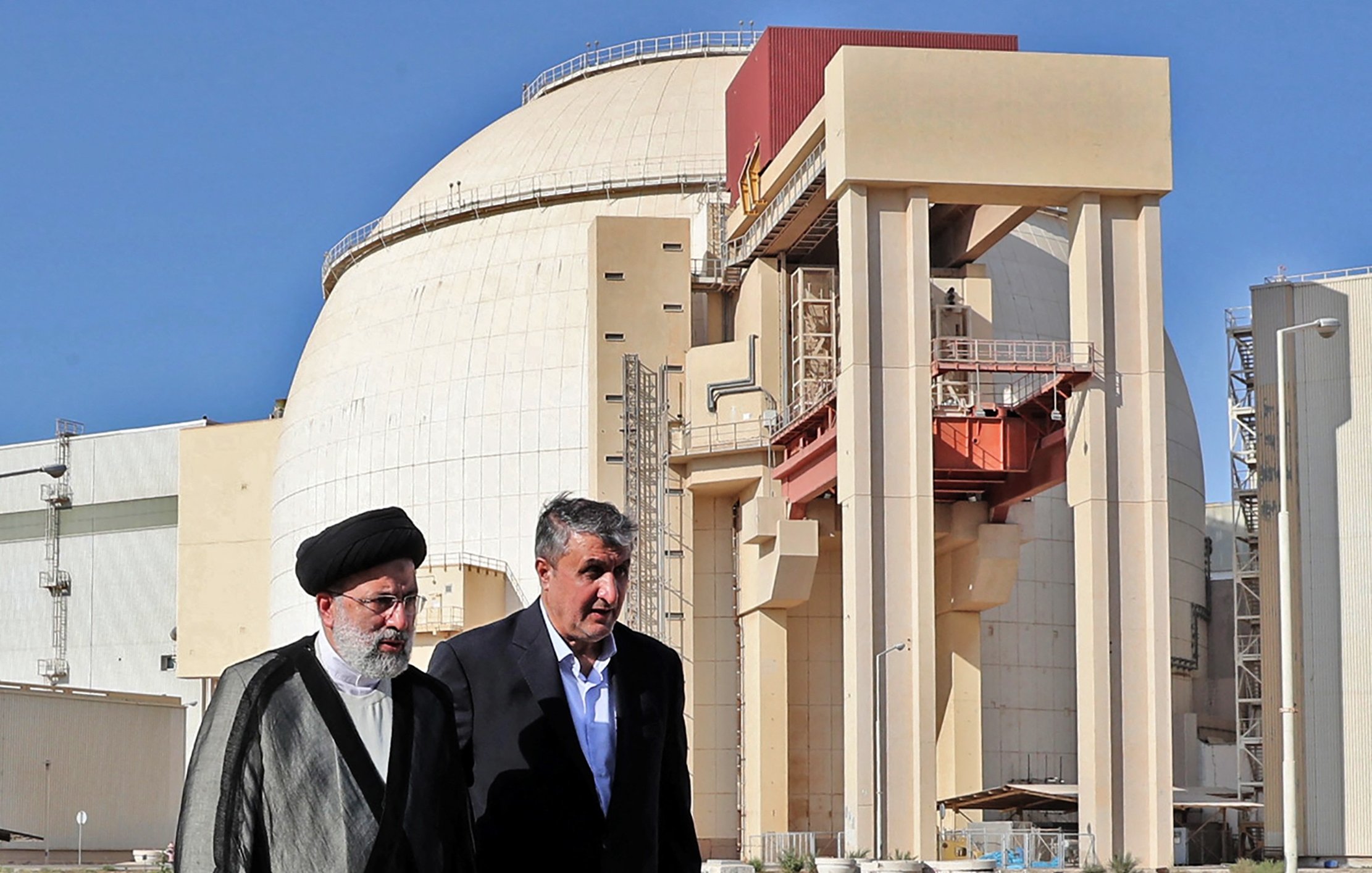 L'image montre deux hommes marchant devant une grande structure industrielle, qui semble être un réacteur nucléaire. Le bâtiment a une forme arrondie et est de couleur claire, avec des éléments de construction en béton visibles. Les deux hommes portent des vêtements formels ; l'un est vêtu d'un costume traditionnel et l'autre porte une veste. L'arrière-plan est dégagé, avec un ciel bleu. Cette scène pourrait illustrer une visite officielle ou une discussion liée à l'énergie nucléaire.
