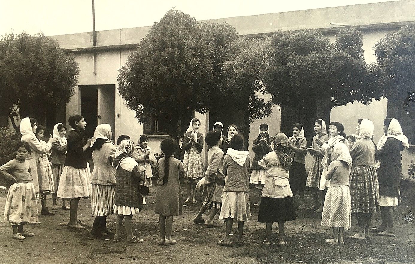 L'image montre un groupe d'enfants, principalement des filles, rassemblés dans une cour. Ils semblent jouer ensemble, entourés de grands arbres. Les filles portent des robes et certaines ont des foulards sur la tête. L'ambiance est joyeuse et festive, suggérant un moment de camaraderie et d'amusement. L'arrière-plan est constitué de murs d'un bâtiment, ce qui donne une atmosphère de communauté.