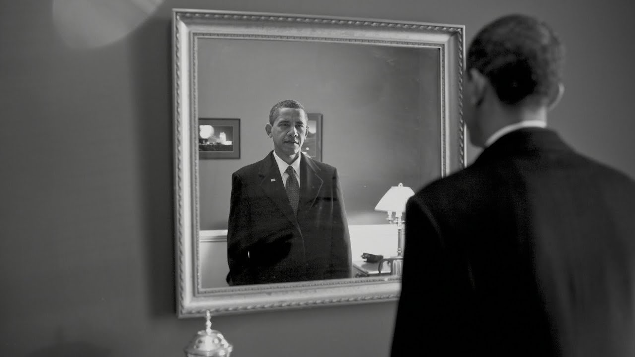 L'image montre un homme en costume noir se tenant devant un miroir. Son reflet dans le miroir affiche une expression sérieuse. L'environnement semble sobre, avec des éléments de décoration tels qu'une lampe et un cadre au mur. La scène est présentée en noir et blanc, ajoutant une atmosphère mélancolique et introspective à l'image.